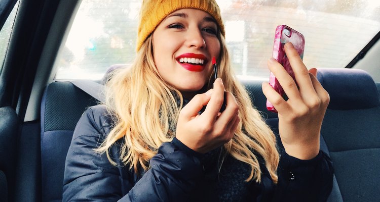 A woman applies makeup in the backseat of a car using her iPhone screen as a mirror.