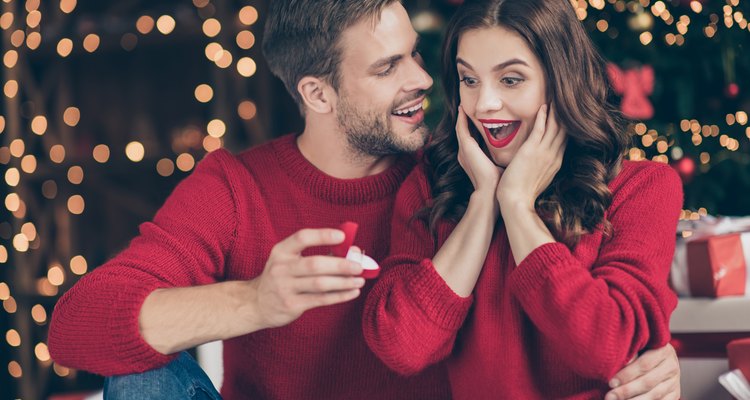 Photo of couple in decorated garland lights room guy giving lady unexpected engage ring box waiting answer sitting cozy near x-mas tree indoors wear red sweaters