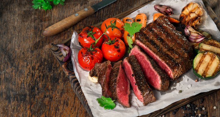 Sliced rare grilled steak on rustic cutting board with set of grilled vegetables on dark rustic wooden table with copy space
