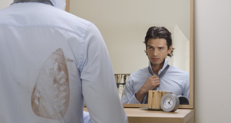 Young man adjusting tie in mirror, iron stain on shirt, rear view