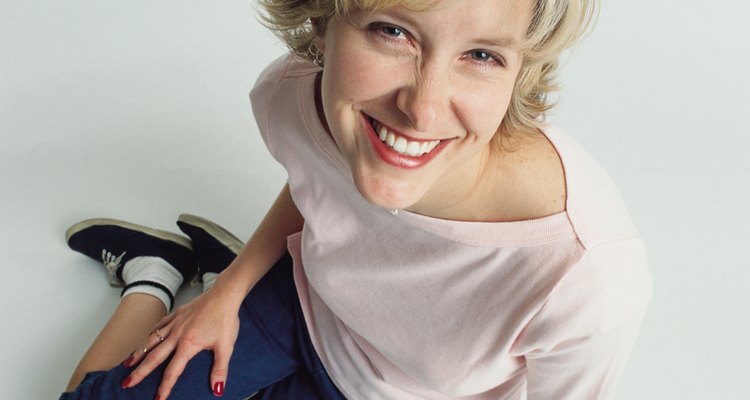 beautiful young slender blonde caucasian adult female wearing jeans and a pale pink t-shirt and tennis shoes sits on the floor as she leans forward smiling at the camera playfully